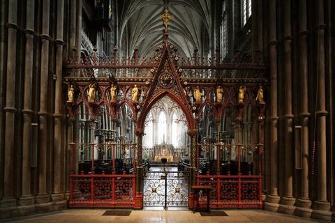 Choir screen. The delicate tracery of a vibrant red and gold screen is backlit from the bright transept beyond. Gold figures line the upper level of the screen. The central opening is a gated archway.