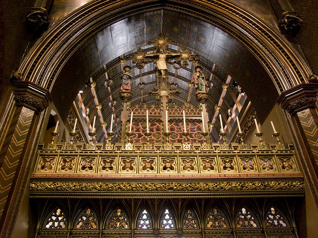 Rood screen. In this colour photograph, we look up at the top zone of a partition that fills an arch in a church. A row of candles lines several bands of gold tracery and ornamental patterns. A crucifix rises into the arch above, and is flanked by two people, the Virgin Mary and St John the Evangelist, standing on miniature platforms to either side.
