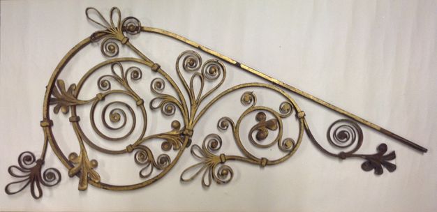 Metalwork. Gold-coloured iron is curved and curled into tightly spiralling swirls and leafy forms. The piece is shaped like a teardrop on its side, with the pointy end to our right.