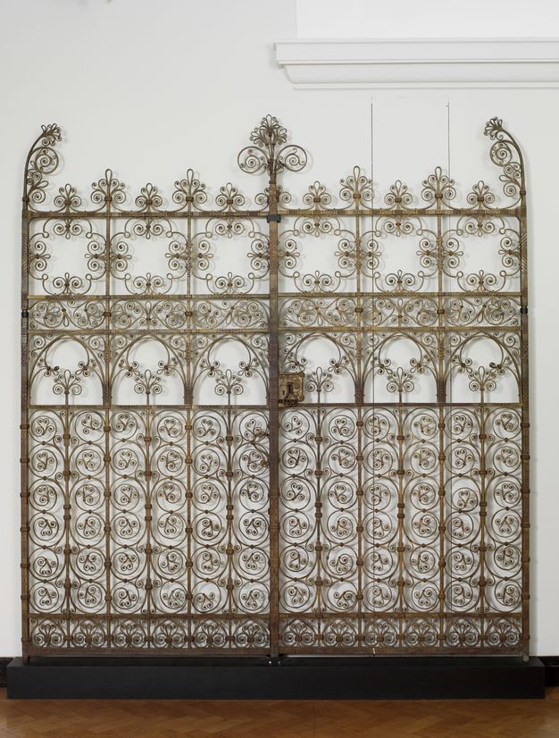 Gates. Gilded wrought iron gates curl and swirl in an intricate pattern reminiscent of filigree. The shadows the gate casts on the white wall behind it make it appear blurry, like double vision.