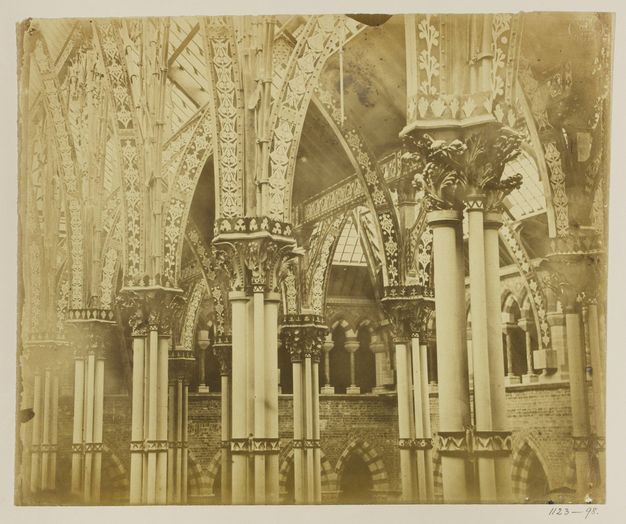 Architecture. Printed in sepia-brown tones, this photograph shows columns holding up heavily ornamented capitals and arches. Additional pointed arches are set into brick walls in the space beyond.