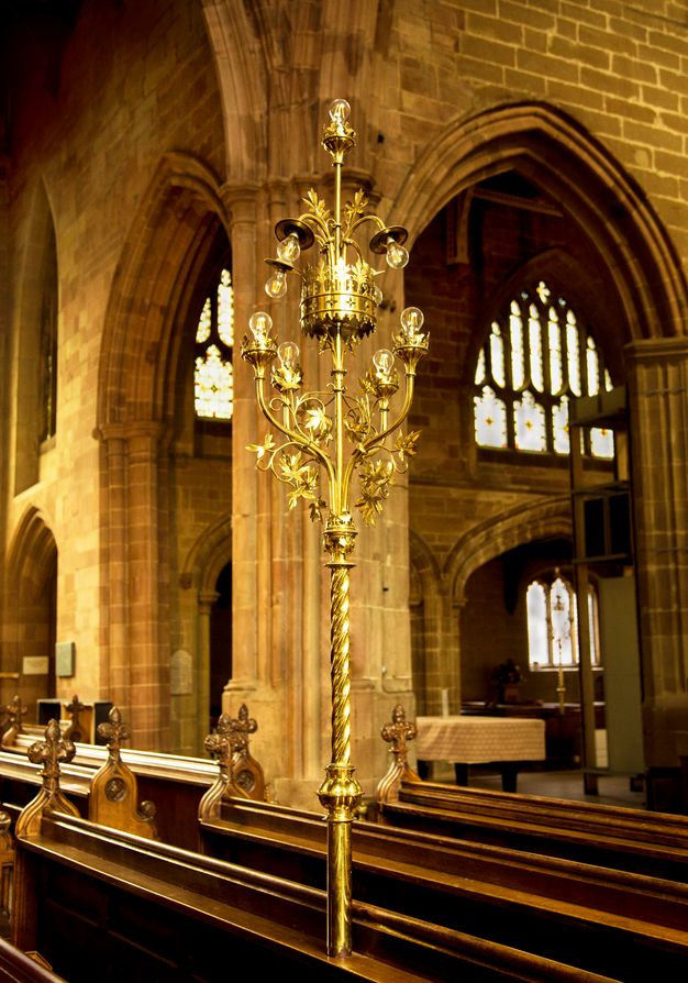 Metalwork. Installed so it rises from the back of a pew, a shiny gold light has two sets of electric bulbs on a tall, twisted shaft. Arms holding the naked bulbs reach out and up like a chandelier, to meet a few more arms bowing down around a crown-like form.