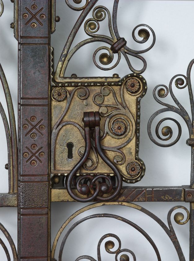 Metalwork. Detail view of a gold, boxy rim lock decorated with swirling vines. The iron pull for the gate is hinged so it hangs down. Two curling strips of iron interlock to make a double-lobed design.