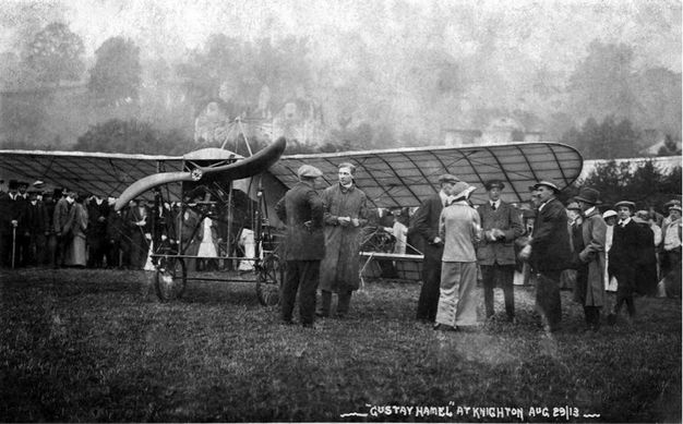 Black and white photograph. Men and women mill around a monoplane with a shallowly curving S-shaped propeller, parked in a grass field.  