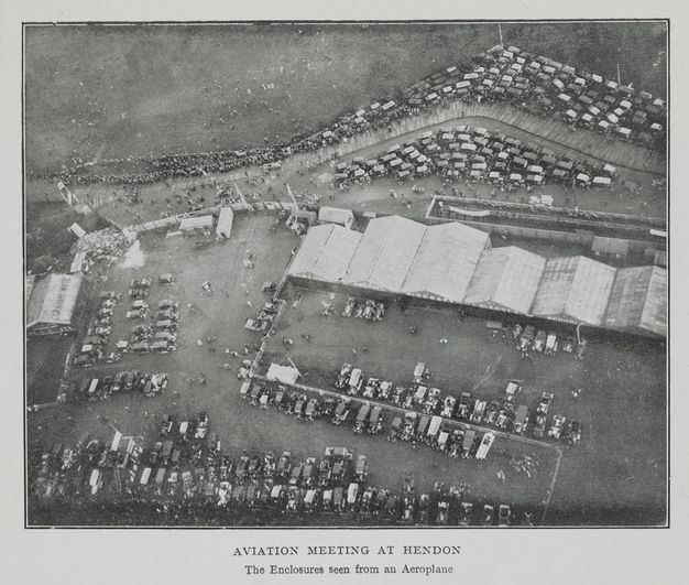 Black and white photograph. A high, aerial view of flat grounds. Six wide hangars are lined up near the centre of the composition. Cars are parked around the edges of the fields, and people dot the landscape. Text below reads, “Aviation Meeting at Hendon The Enclosures seen from an Aeroplane”.