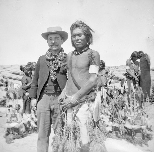 Black and white photograph. A pale skinned man wearing a suit stands smiling next to a brown-skinned man wearing Hopi dress. Both look at the camera. The Hopi man is bare-chested, has dark, chin-length hair, and his lips are closed. More Hopi Indians stand in a desert in the background.