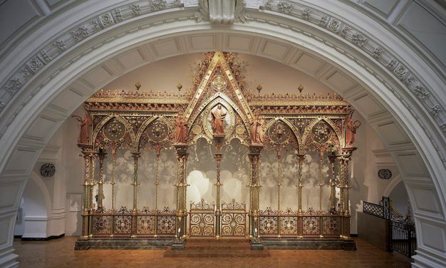 Choir screen. This colour photograph shows a freestanding architectural partition, like a floating wall, has five bays, each under a pointed arch. Each arch is then bisected by a spindly column. The central bay is the widest and tallest, and a person stands at its apex. Intricate tracery encrusts the upper edges of the screen and fills in the arches. Panels across the bottom are also made with intricately patterned tracery. Most surfaces are shiny or burnished gold, and there are also touches of muted red, warm yellow, and earthy green.