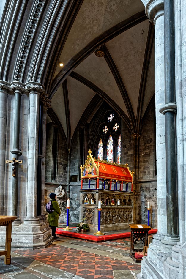 Colour photograph. A rectangular stone tomb is topped with a gallery-like level of pointed arches, which is under a canopy painted vivid red and blue. The triangular front face of the canopy is painted with a scene of the Virgin Mary with saints and angels, who hold up a map. A person standing in the church in front of the shrine in this image is about half the height of the whole shrine.