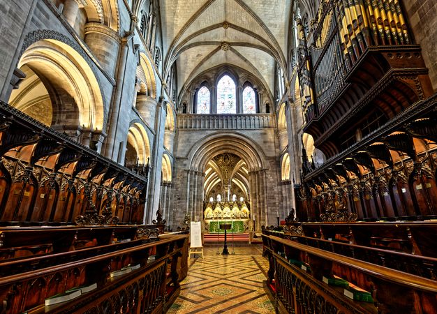 Architecture. Colour photograph of a narrow aisle flanked on both sides by rows of continuous wooden seats. The three bays of the choir and vaulted ceiling soar overhead. Three stained glass windows are set into far wall of the transept in front of us.