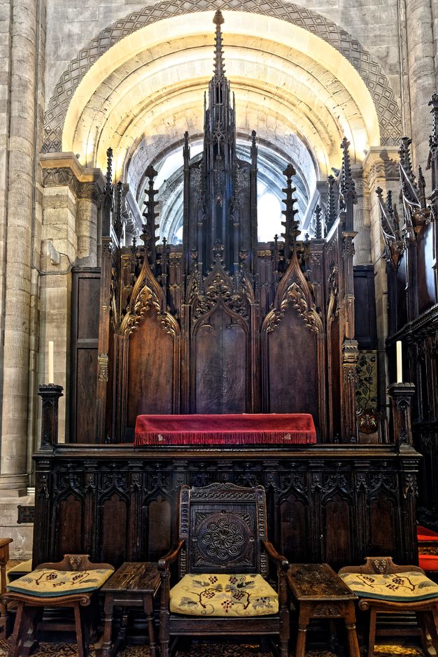 Architecture. Colour photograph of a wide, carved wooden throne that sits three across, each under a pointed arch carved with tracery and spiky spires above. A panel, also intricately carved, cordons off the space of the throne, like a footboard of a sleigh bed. Three more chairs with seats upholstered in fabric patterned with grape vines against a butter-yellow background are in front of the throne.