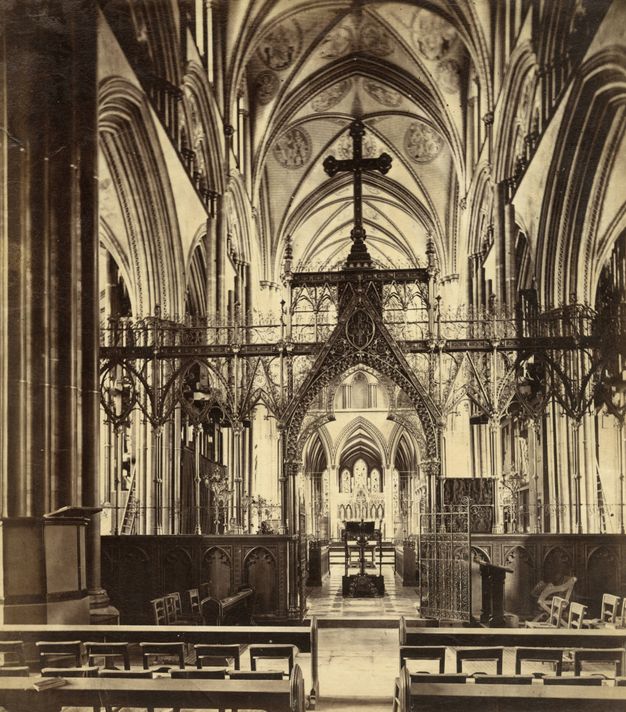 Choir screen. Black and white photograph of a lacy, almost entirely see-through choir screen crosses the clustered columns and immense vaults of the nave. The tracery is so delicate it looks impossibly light.