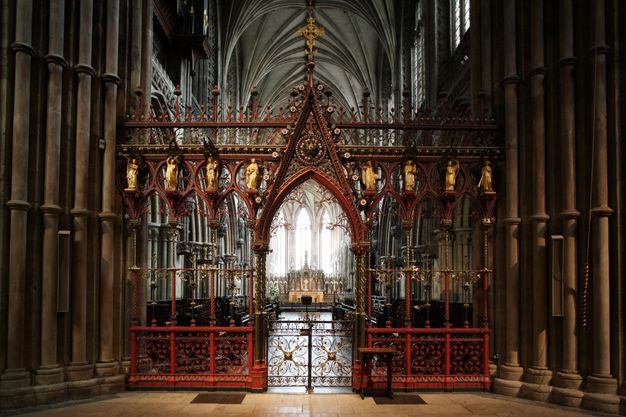 Choir screen. Colour photograph in which the delicate tracery of a red and gold screen is backlit from the bright transept beyond. Gold figures line the upper level of the screen. The central opening is a gated archway.